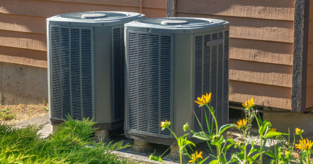 Two HVAC systems placed outside a home surrounded by grass and yellow flowers