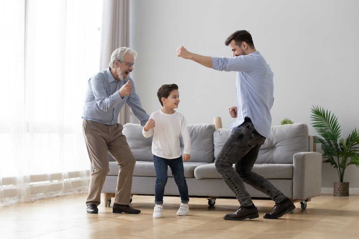 Happy grandfather, father and little son having fun at home, excited granddad, dad and preschool child boy grandchild dancing to favorite music together, family playing in living room, funny activity